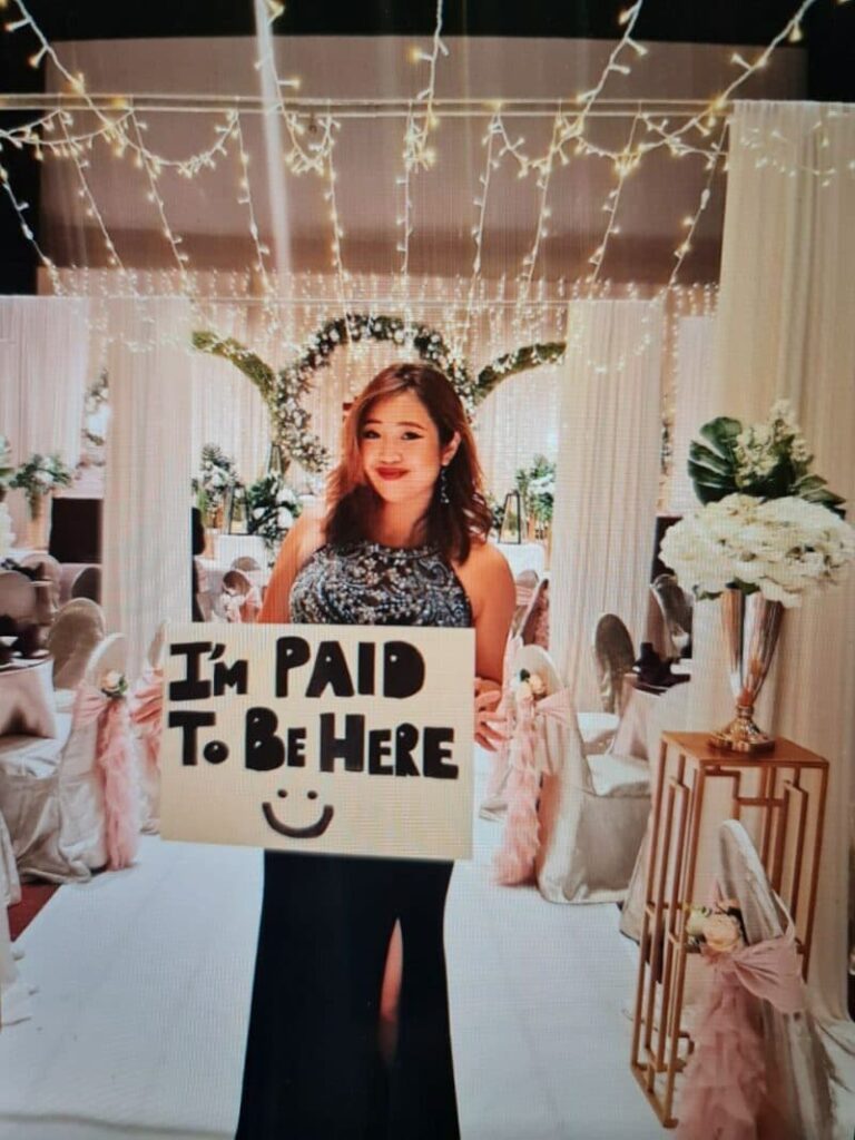 A portrait of Jemma holding a sign saying "I'm Paid To Be Here"